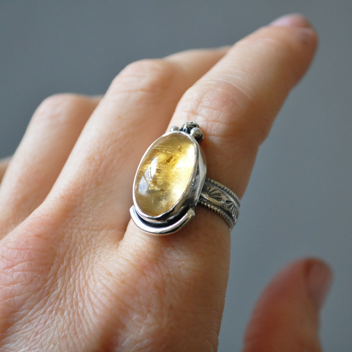 A silver ring with a large oval yellow citrine gemstone, worn on a person's finger against a soft gray background.