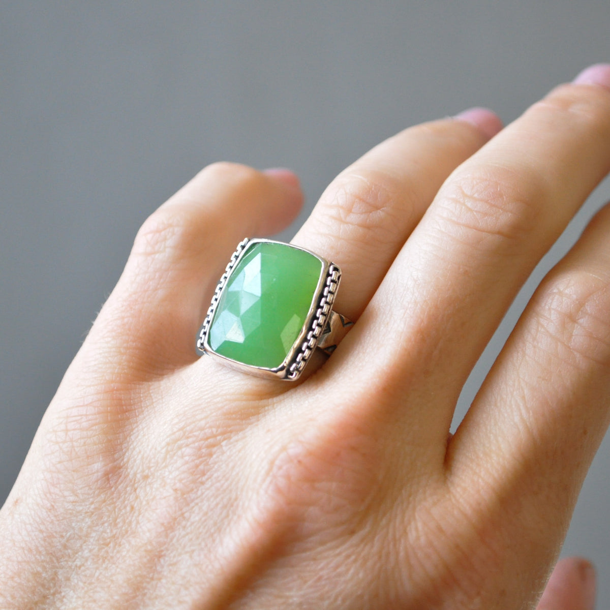 A silver ring with a large, cushion-cut green chrysoprase stone featuring a faceted center, worn on a person's finger against a neutral gray background.