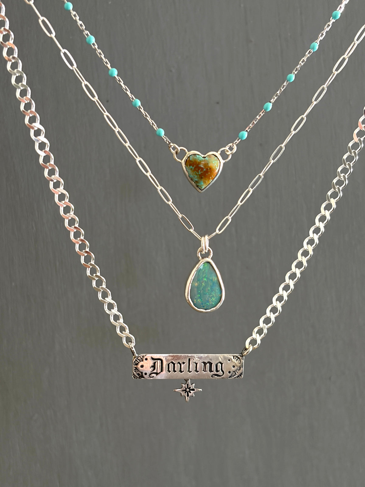 Three layered silver necklaces displayed against a dark gray background. The top necklace features small turquoise beads and a heart-shaped green stone pendant.