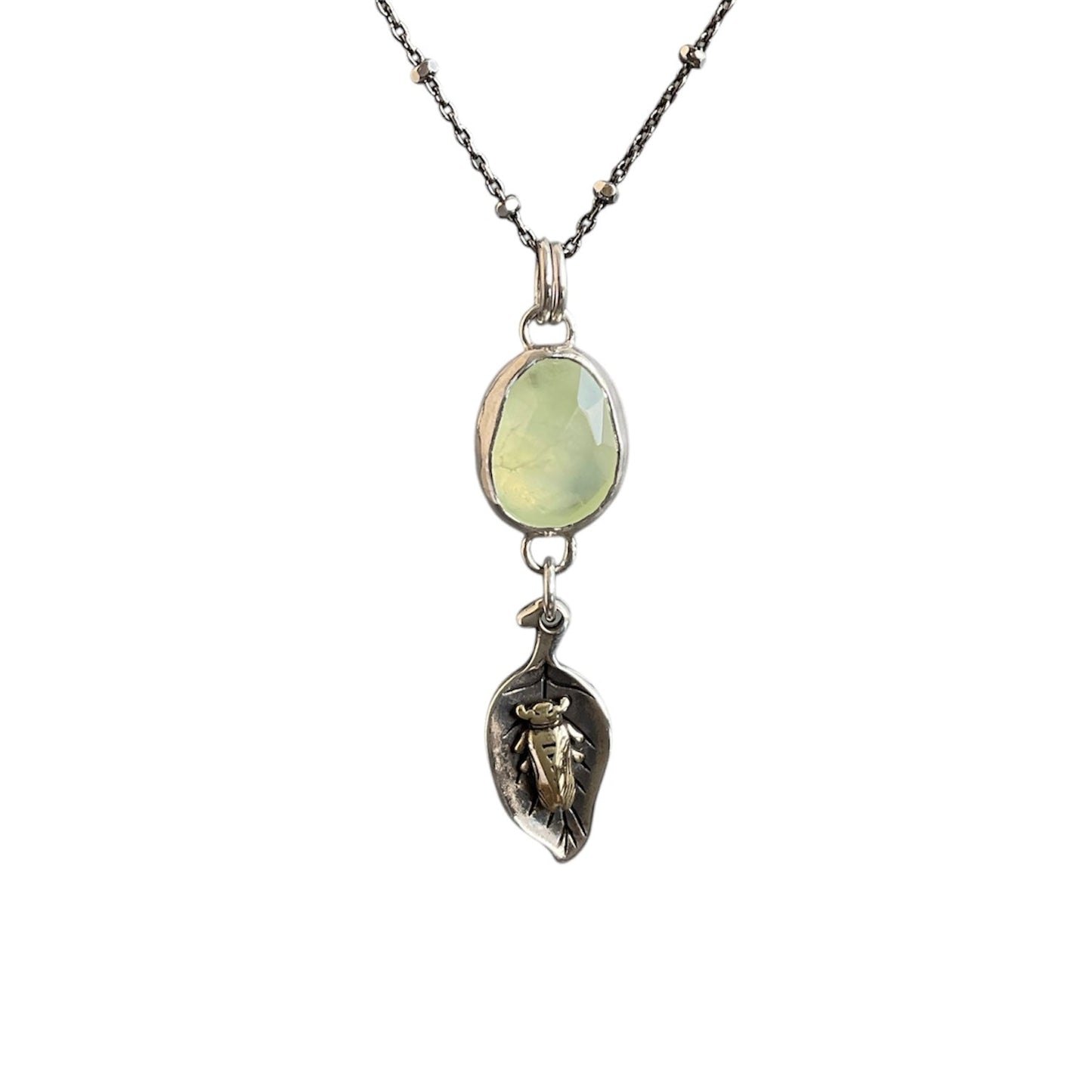 A light green faceted prehnite gemstone bezel set and hung above a leaf charm holding a brass sleepy beetle.
