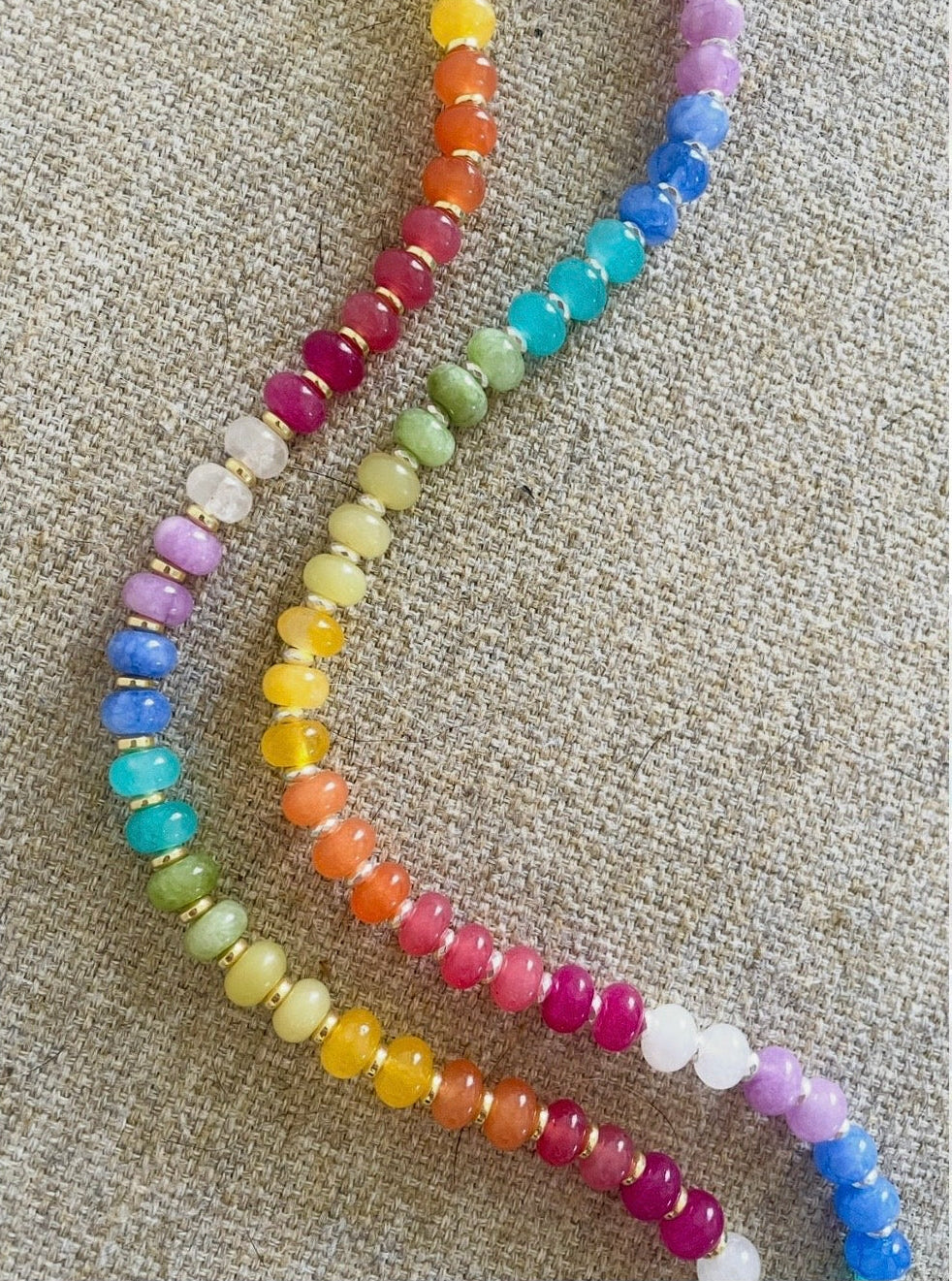 A strand of colorful round beads arranged in two parallel rows on a textured beige fabric background. The beads transition through rainbow colors including pink, orange, yellow, green, blue, and purple.