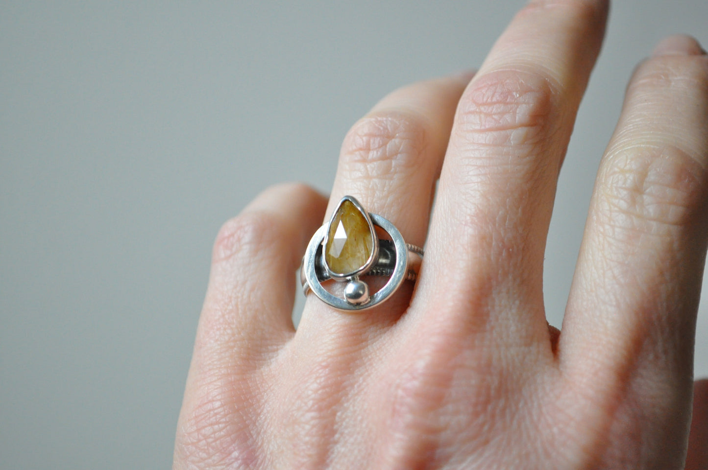 Silver ring with a yellow gemstone on a person's finger against a neutral background