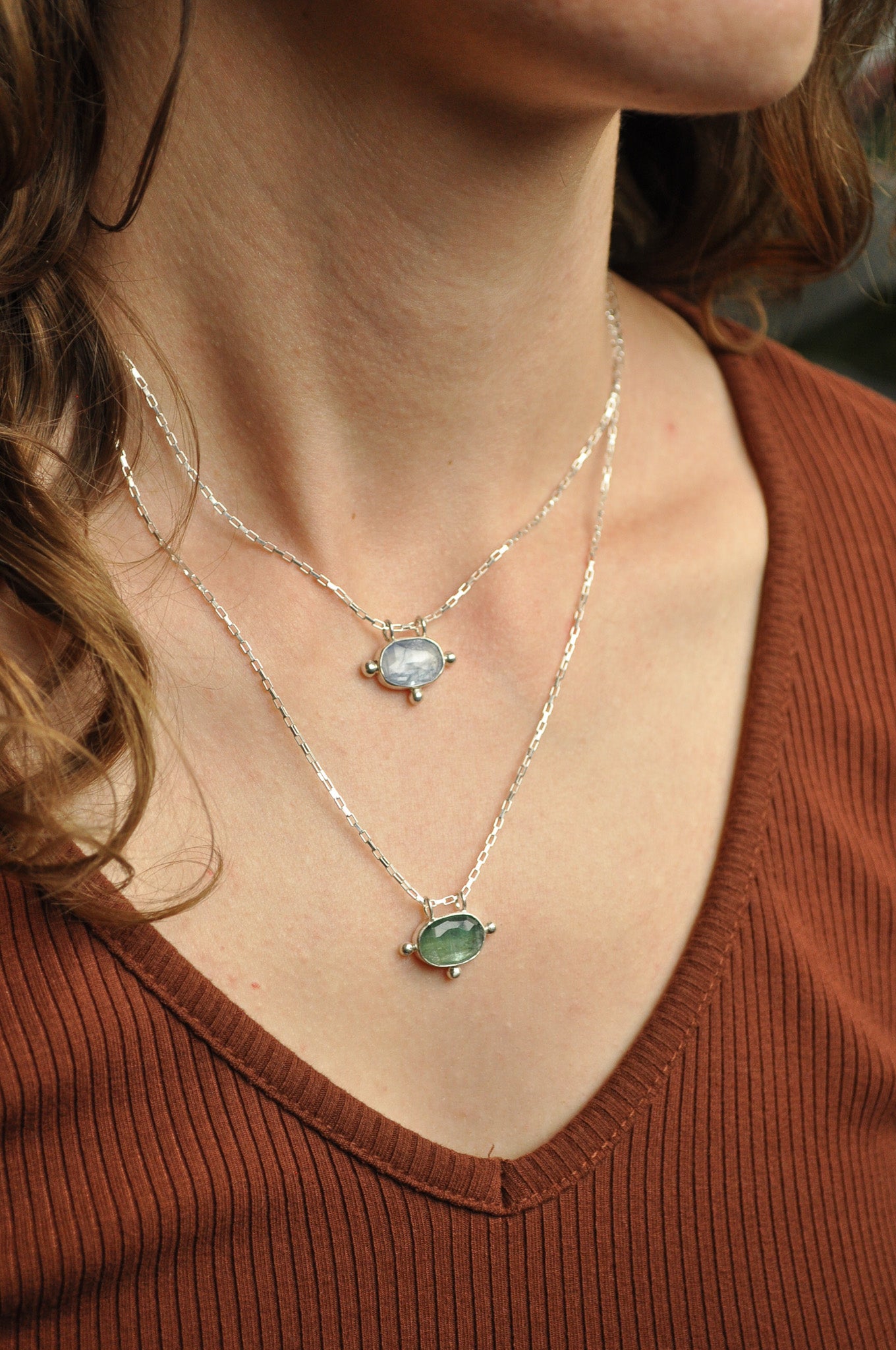 Close-up of a person wearing two necklaces with gemstones on a brown shirt.