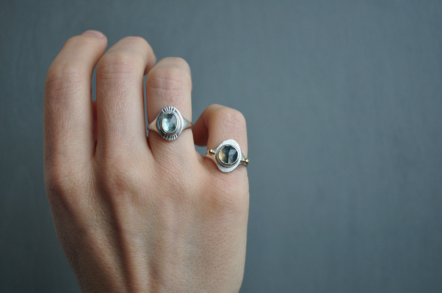 Hand wearing two silver rings with gemstones against a gray background