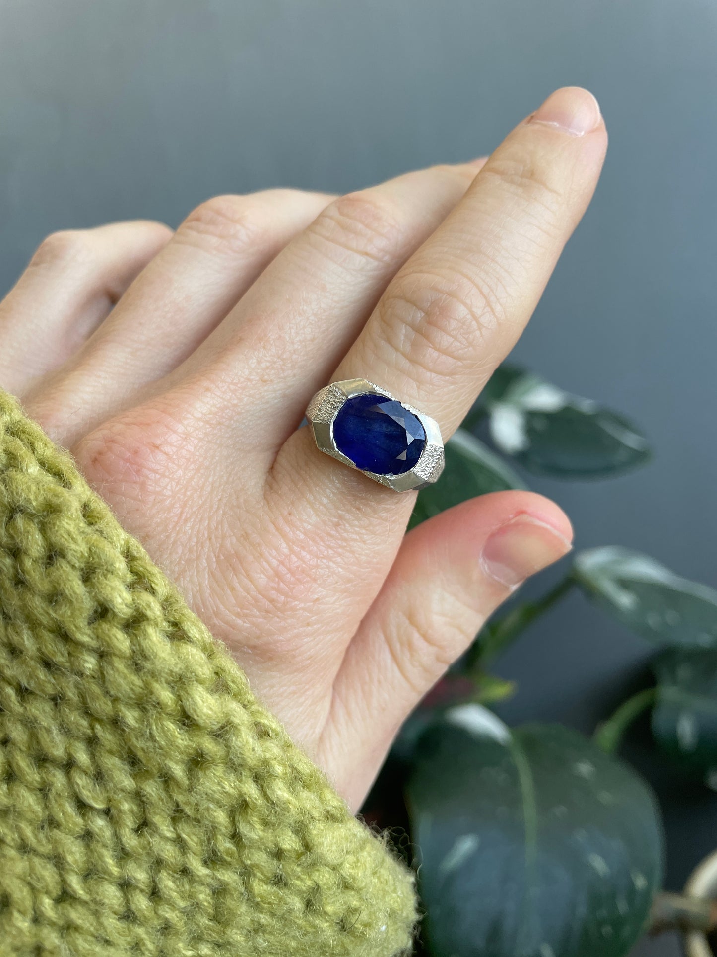 Hand wearing a ring with a blue gemstone, blurred green leaves in the background