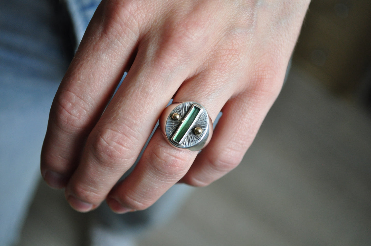 Close-up of a hand wearing a silver ring with a green stone on a blurred background