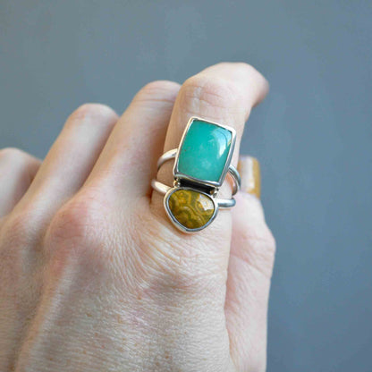 Hand wearing a ring with green and yellow gemstones on a gray background