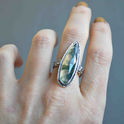 Hand wearing a ring with a large, teardrop-shaped gemstone on a neutral background