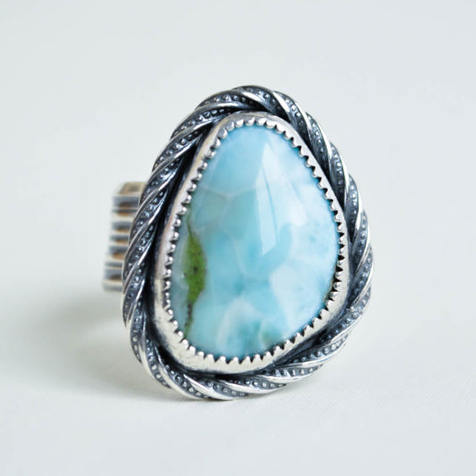 A light blue and white smooth larimar cabochon set in sterling silver with a twist patterned border and a wide ridged band.