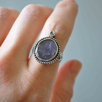 Silver ring with a purple stone on a person's finger against a neutral background