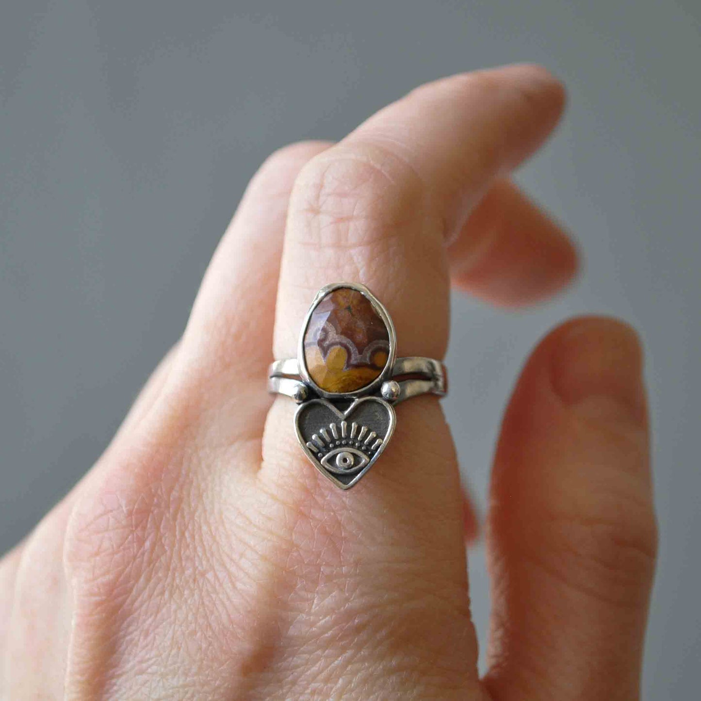 Hand wearing a silver ring with a heart and eye design on a neutral background