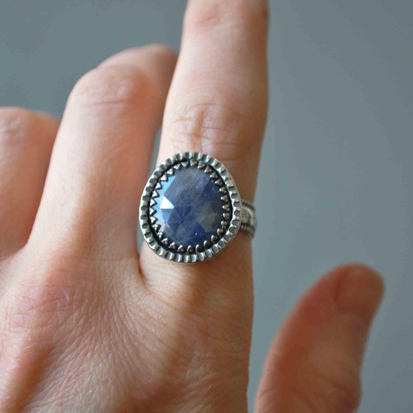 Silver ring with a blue gemstone on a finger against a neutral background
