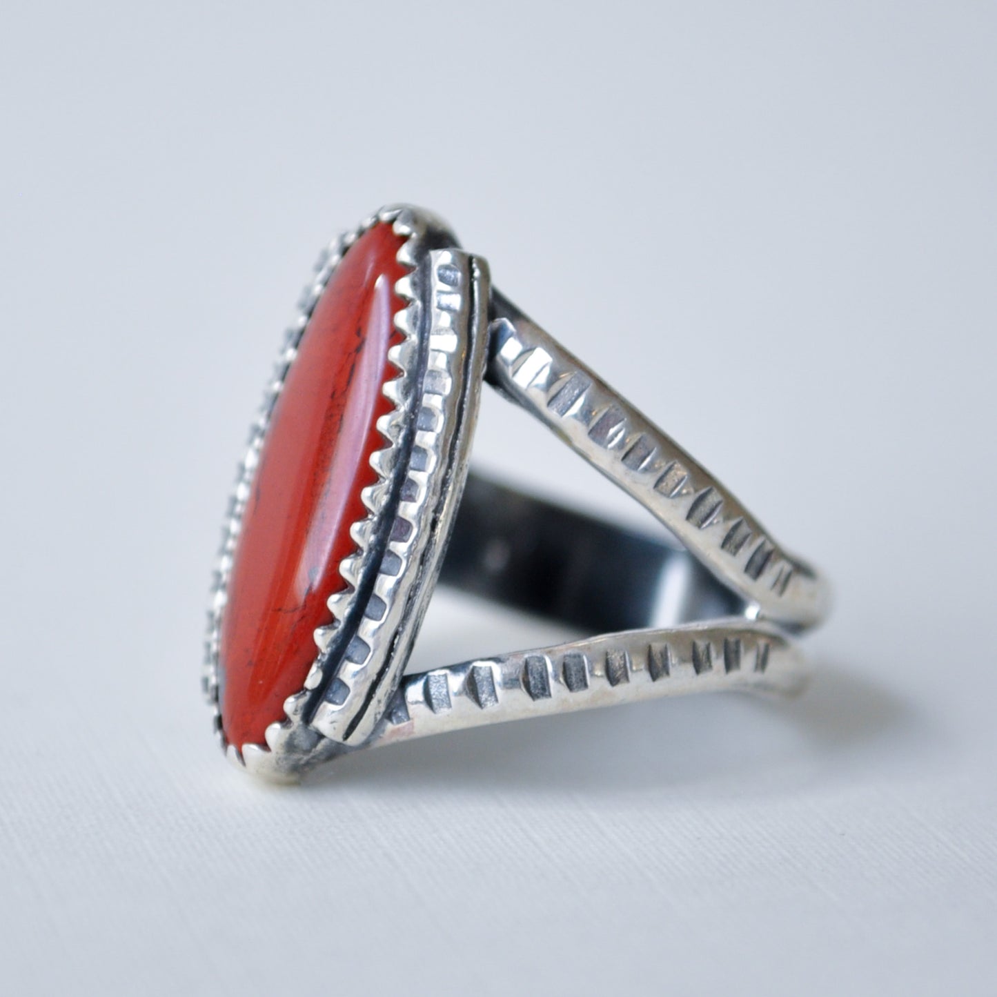 A sterling silver ring featuring a large, oval-shaped red jasper cabochon stone, photographed on a clean, light gray textured surface.