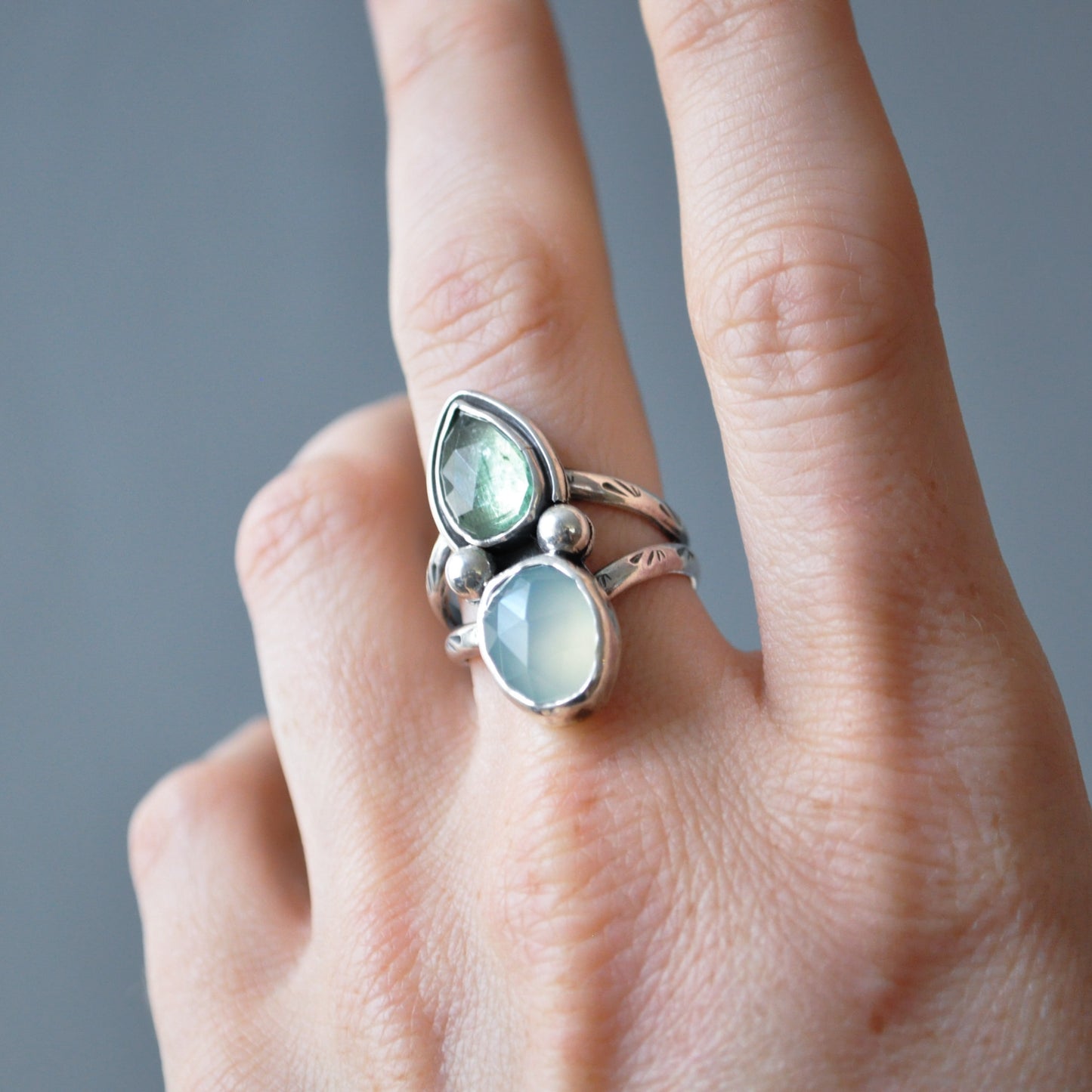 A close-up of a hand wearing a silver ring with two light green stones, one teardrop-shaped and one oval, set against a soft gray background.