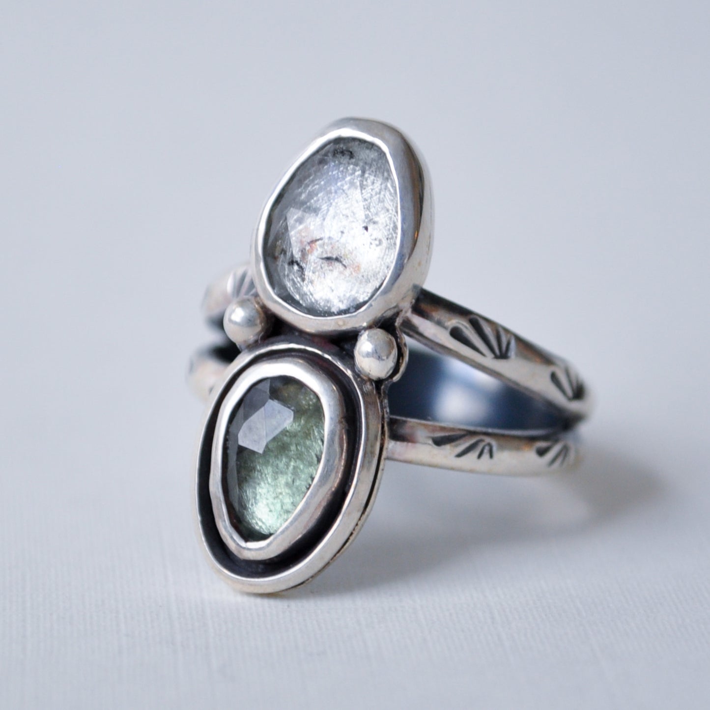 A sterling silver ring with two oval gemstone settings, one clear and one light green, accented with small white pearls, on a light gray background.