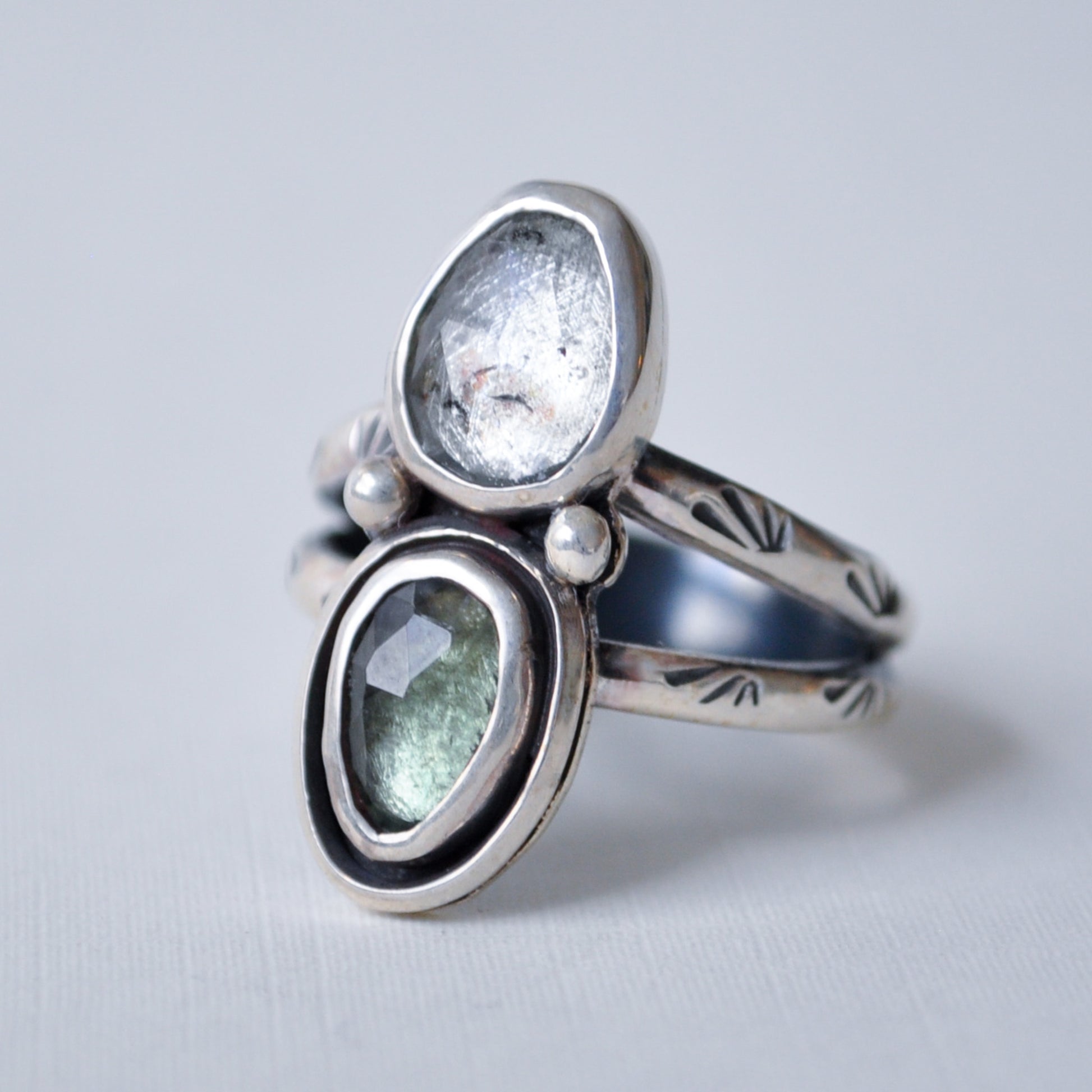 A sterling silver ring with two oval gemstone settings, one clear and one light green, accented with small white pearls, on a light gray background.