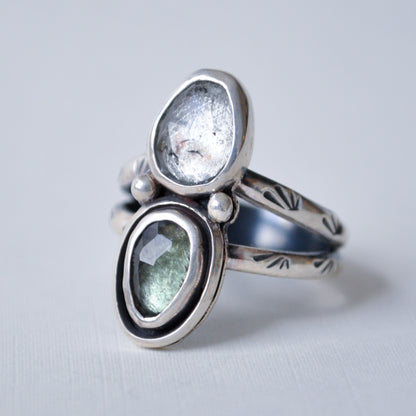 A sterling silver ring with two oval gemstone settings, one clear and one light green, accented with small white pearls, on a light gray background.