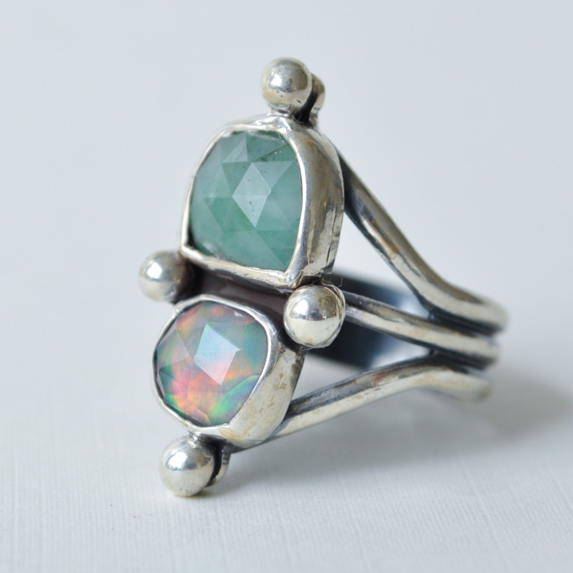 A sterling silver ring featuring two faceted gemstones, one light green and one iridescent opal with rainbow reflections, set in a split-band design on a light gray background.