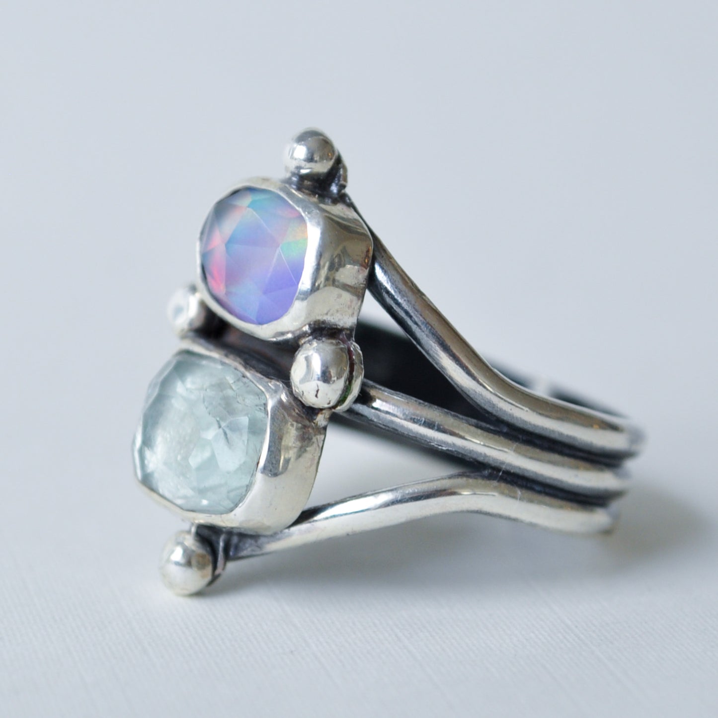 A sterling silver ring with two bezel-set stones, one iridescent purple-blue cabochon and one pale blue-green rough-cut stone, shown on a light gray background.