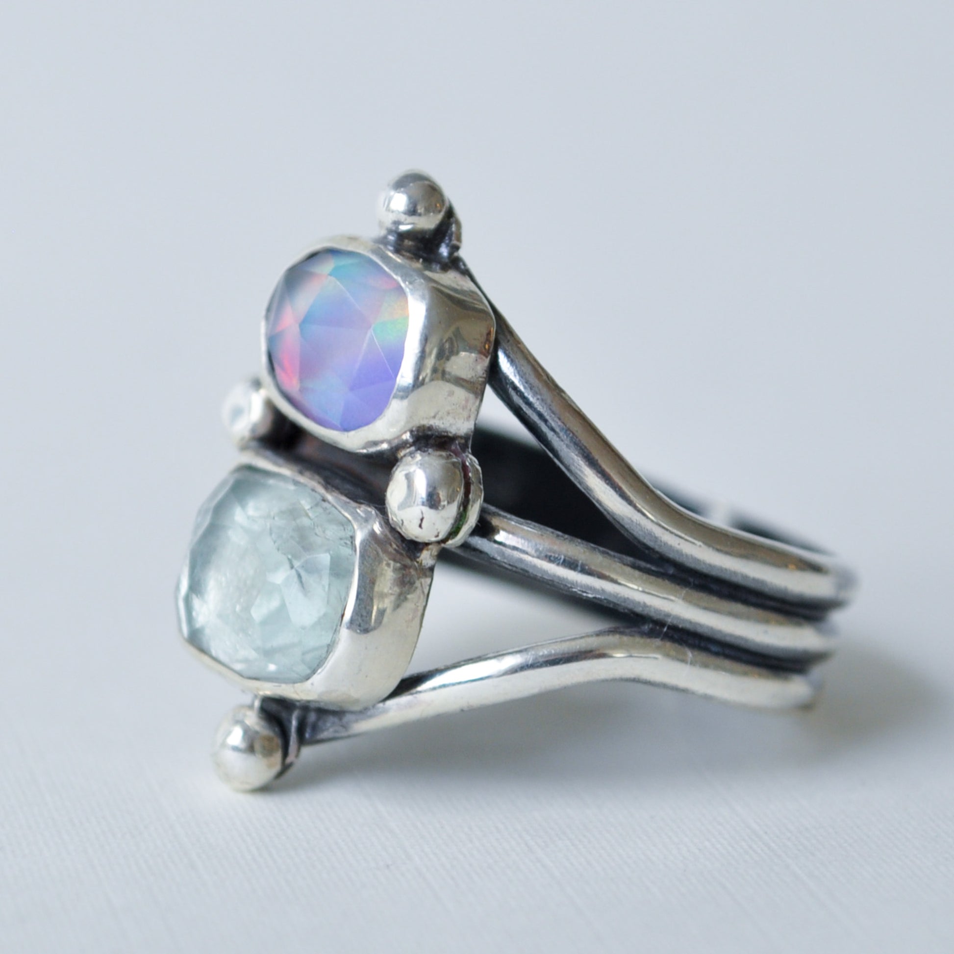 A sterling silver ring with two bezel-set stones, one iridescent purple-blue cabochon and one pale blue-green rough-cut stone, shown on a light gray background.
