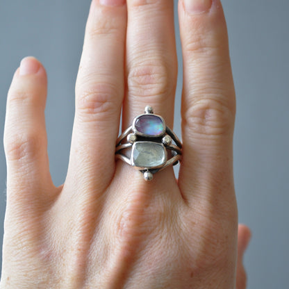 A sterling silver ring with two iridescent stones, one blue and one green, worn on a hand against a soft gray background.