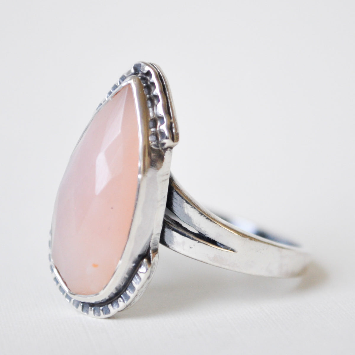 A silver ring featuring a large, teardrop-shaped pale pink opal gemstone in a textured bezel setting, photographed on a clean white background.