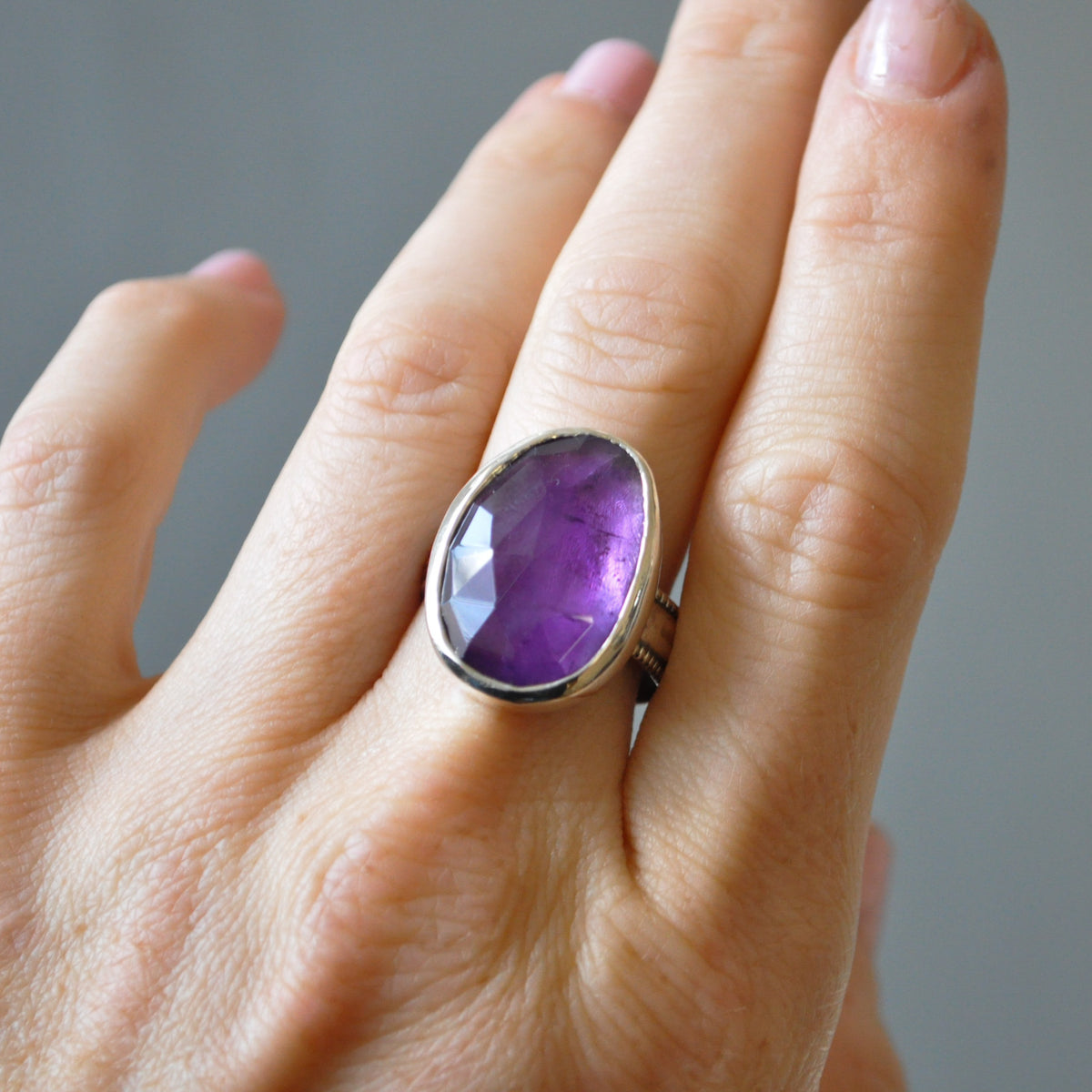 A close-up of a hand wearing a silver ring with a large, faceted oval purple amethyst gemstone.