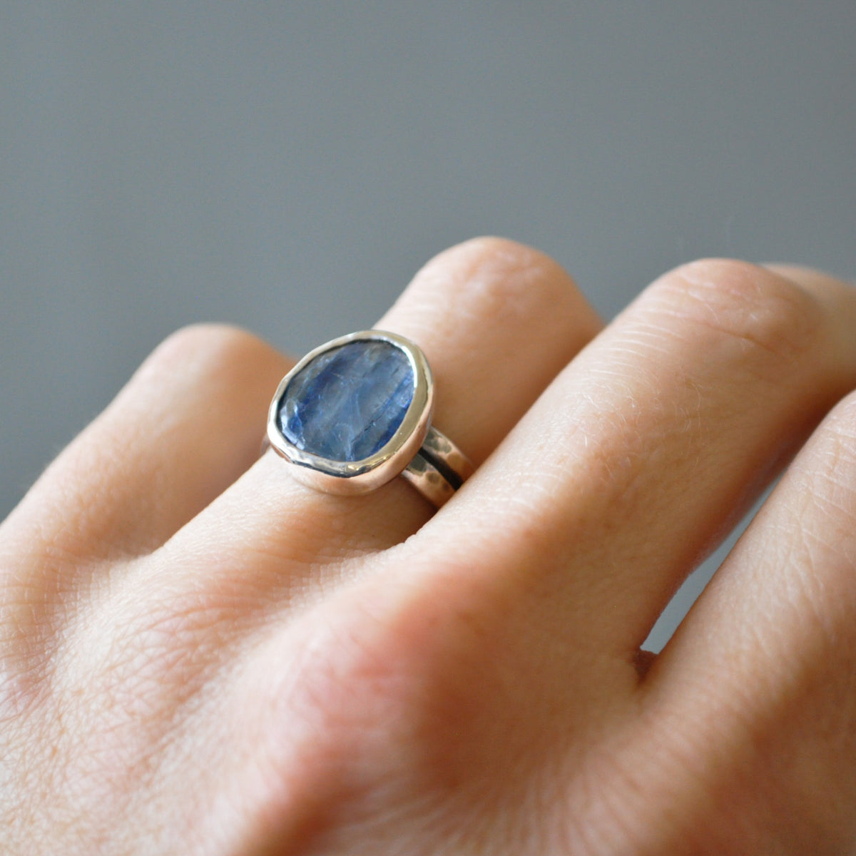A silver ring with a large, irregularly shaped blue kyanite stone, worn on a person's finger against a soft gray background.