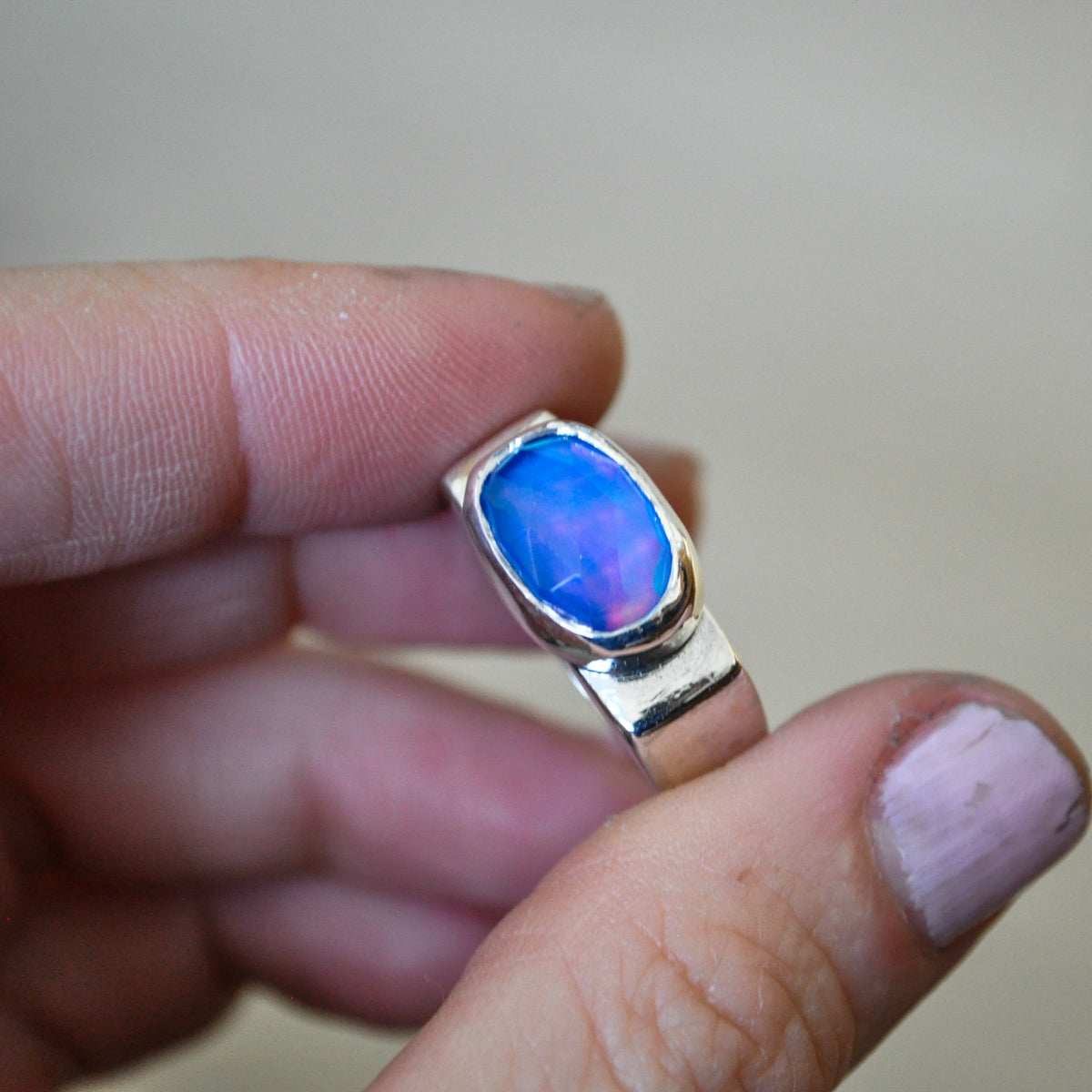 A hand holding a silver ring with a vibrant blue and purple iridescent opal gemstone, set against a soft beige background.