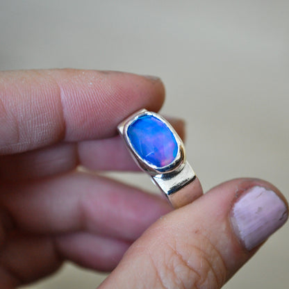 A hand holding a silver ring with a vibrant blue and purple iridescent opal gemstone, set against a soft beige background.