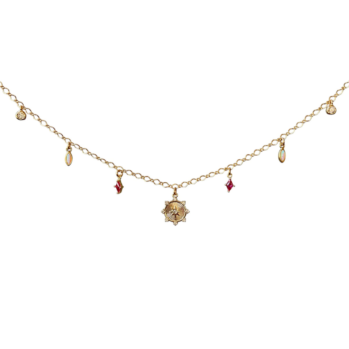 A delicate gold chain necklace featuring a central sunburst pendant and small dangling charms, including red gemstone accents and oval beads, displayed on a clean white background.
