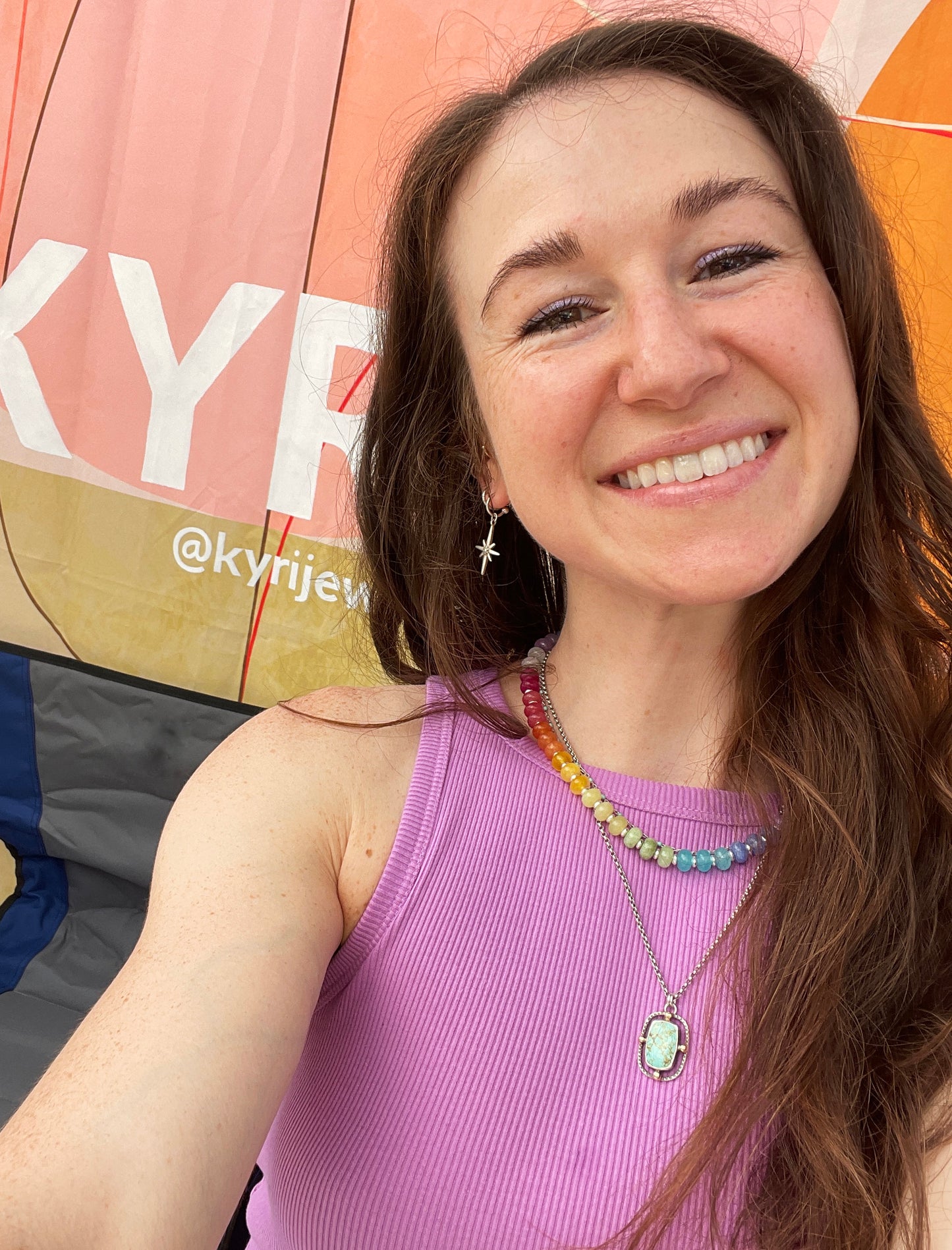 A smiling woman with long brown hair takes a selfie in front of a pink and orange tent. She is wearing a ribbed lavender sleeveless top and layered necklaces, including a silver star earring.