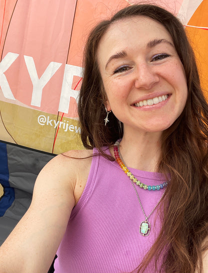 A smiling woman with long brown hair takes a selfie in front of a pink and orange tent. She is wearing a ribbed lavender sleeveless top and layered necklaces, including a silver star earring.