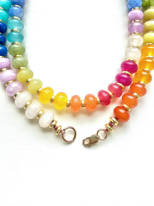 A close-up of a beaded necklace with multiple strands of smooth, polished round beads in rainbow colors separated by small gold spacer beads, with a lobster clasp closure on a white background.