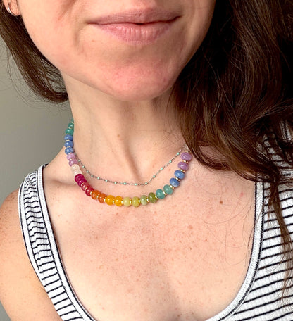 A close-up of a woman wearing a double-strand beaded necklace with a rainbow gradient of translucent beads and a silver chain layered underneath.