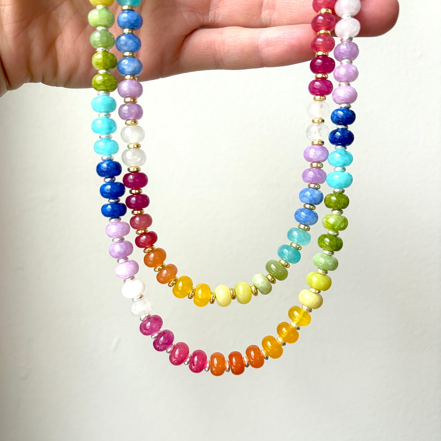 A close-up of a hand holding a long beaded necklace with multiple strands of glossy round beads in rainbow colors separated by small gold spacer beads, against a plain light background.