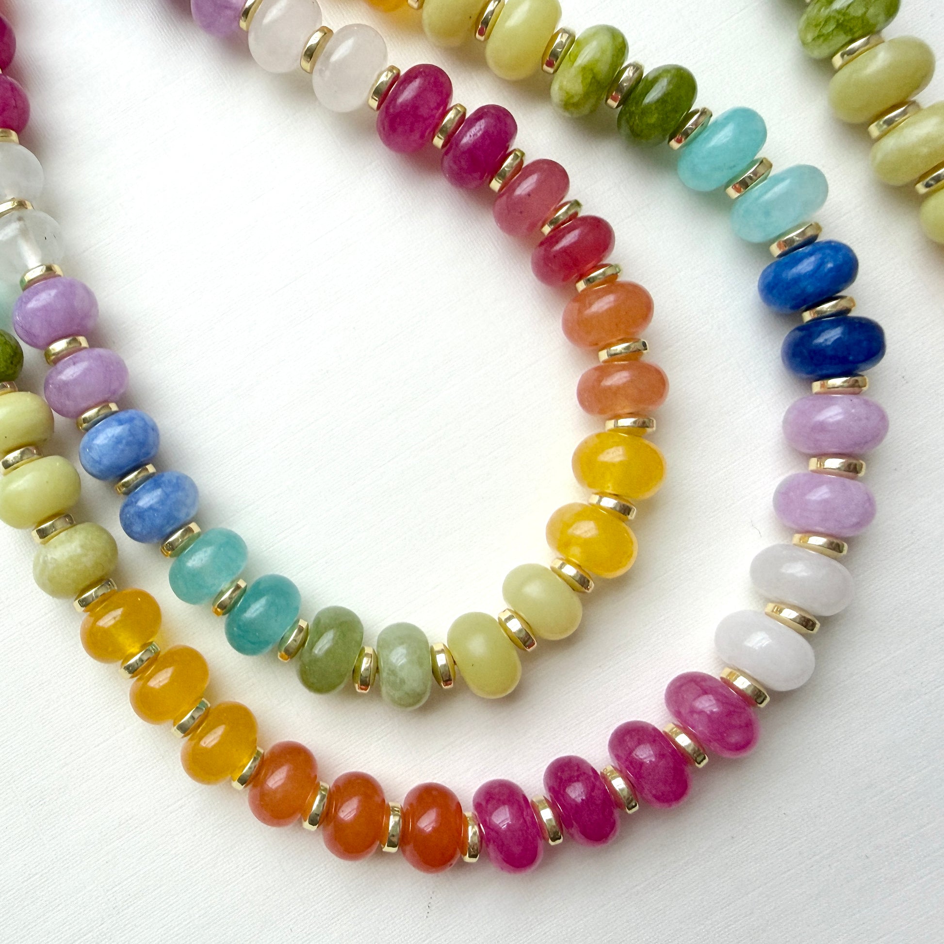 A close-up of a beaded strung necklace featuring multiple strands of colorful round gemstone beads separated by small gold spacer rings, arranged on a white background.