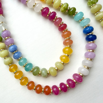 A close-up of a beaded strung necklace featuring multiple strands of colorful round gemstone beads separated by small gold spacer rings, arranged on a white background.