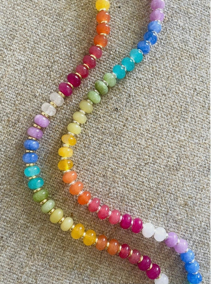 A strand of colorful round beads arranged in two parallel rows on a textured beige fabric background. The beads transition through rainbow colors including pink, orange, yellow, green, blue, and purple.