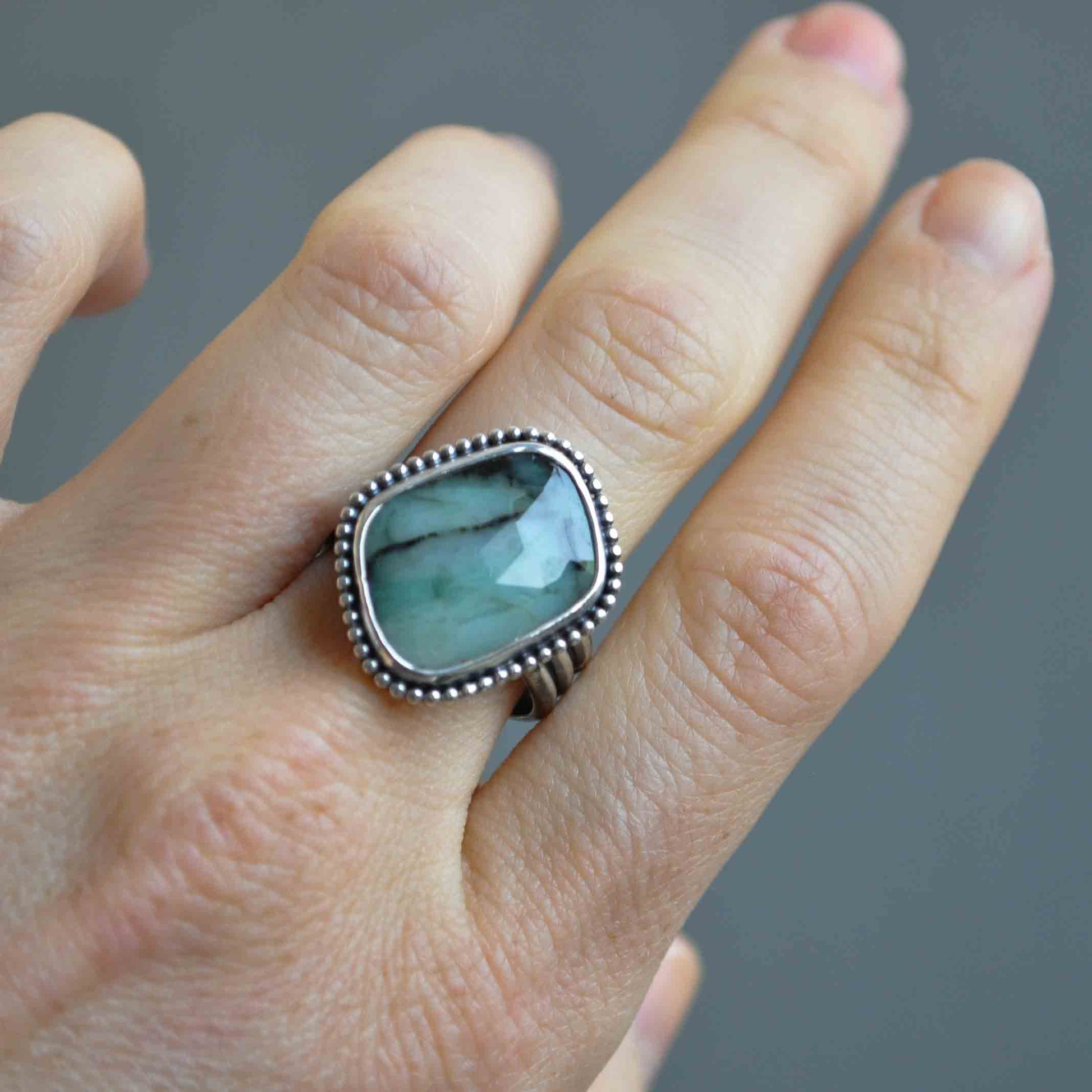 Silver ring with an emerald stone on a finger against a gray background