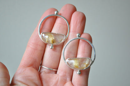Silver earrings with circular design held in a hand against a neutral background