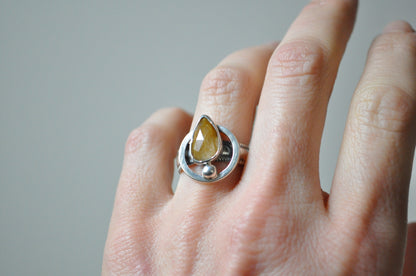 Silver ring with a yellow gemstone on a person's finger against a neutral background