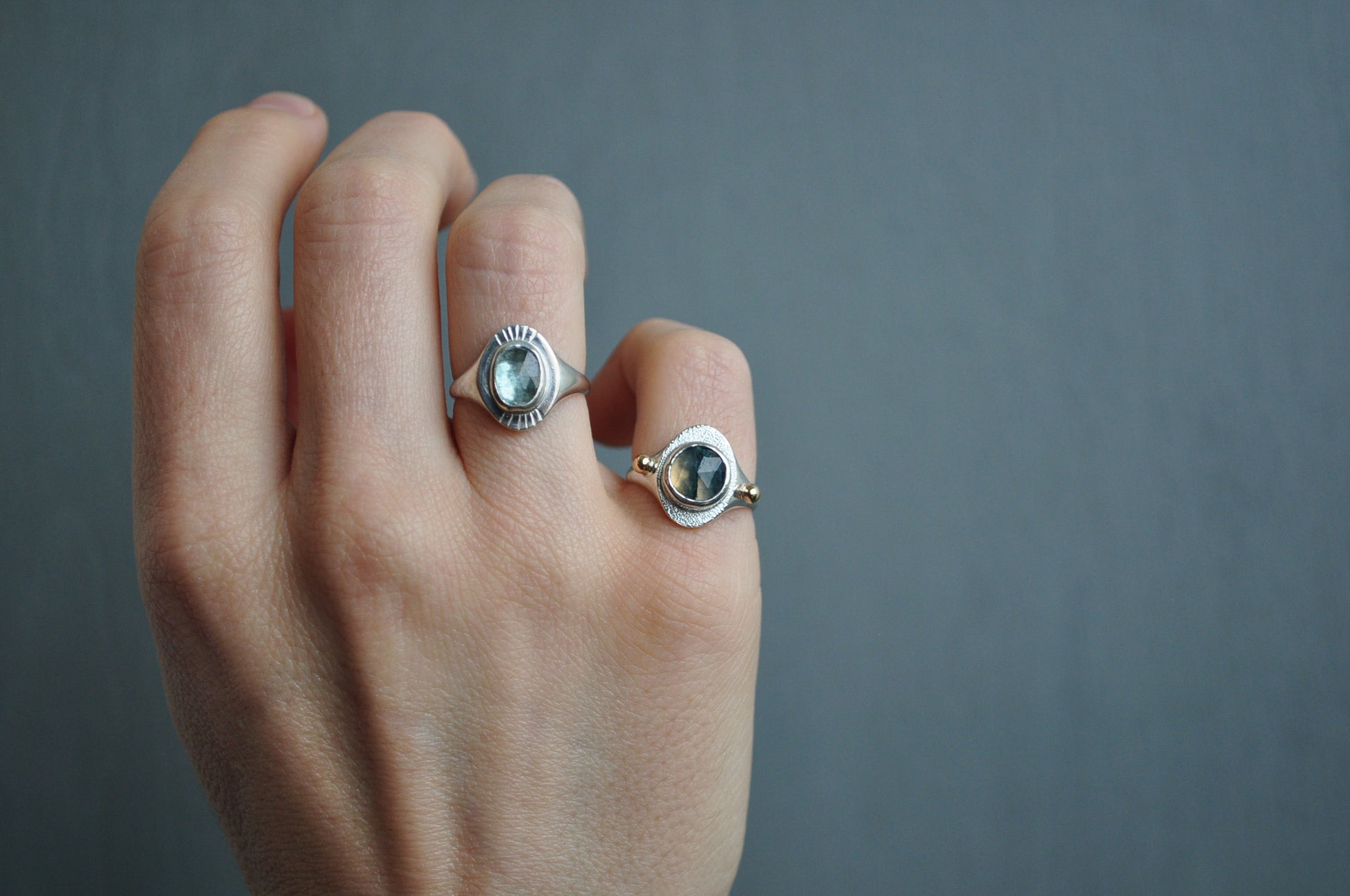 Hand wearing two silver rings with gemstones against a gray background