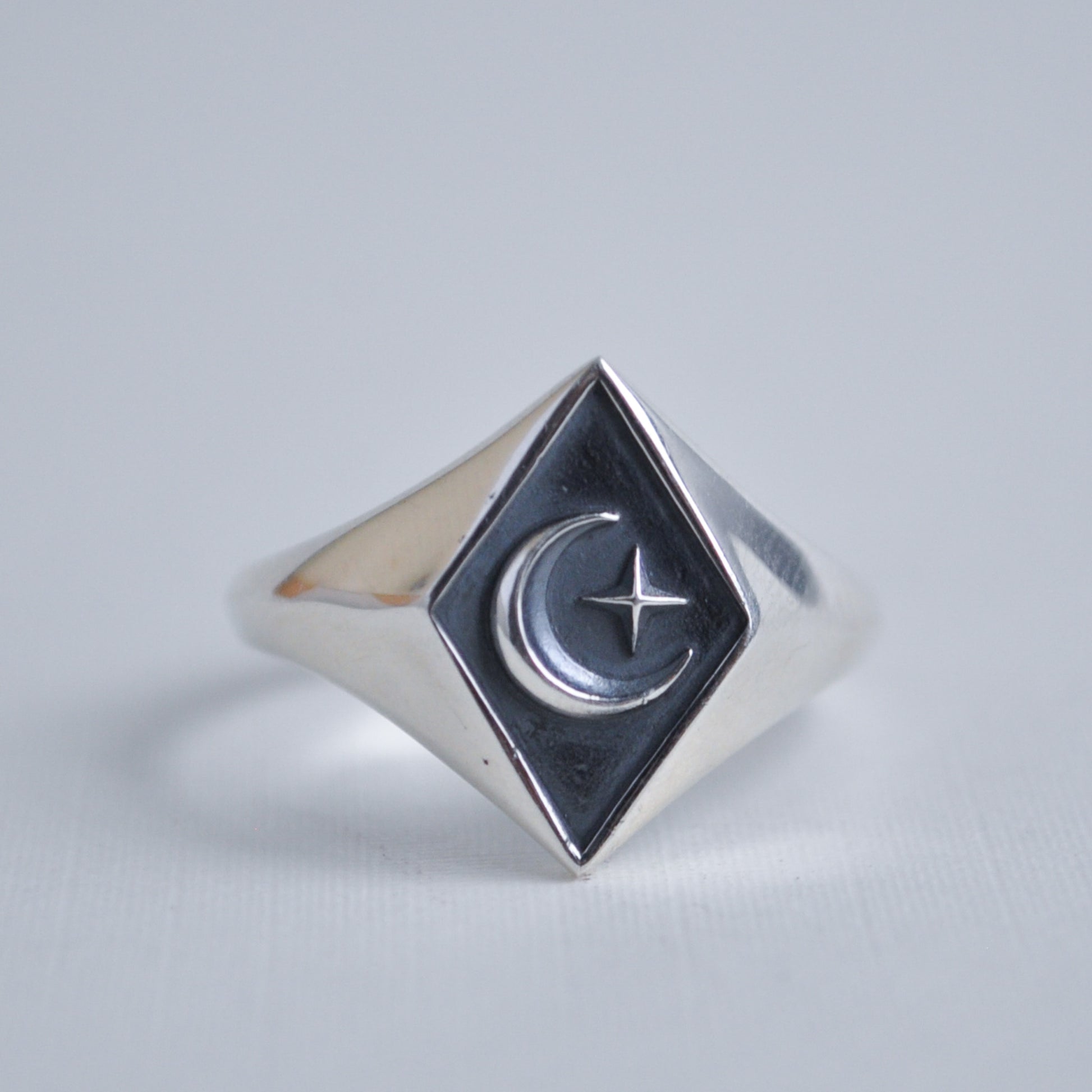 Silver ring with a diamond-shaped face featuring a moon and star design on a light gray background.