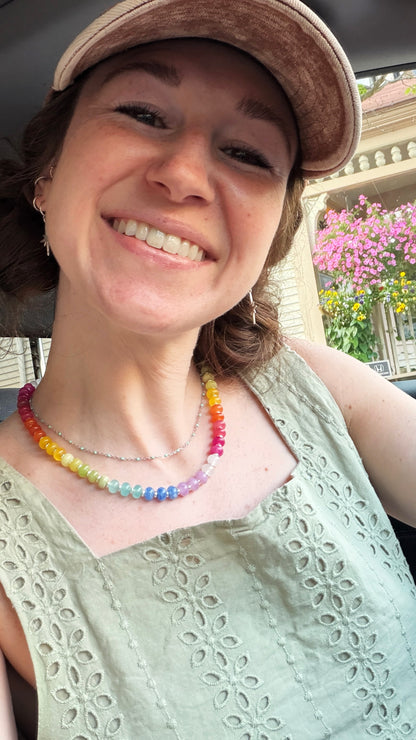 A smiling woman wearing a pastel rainbow beaded necklace and a light green eyelet top, photographed in a casual close-up selfie.