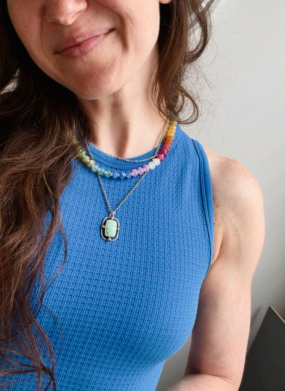 A close-up lifestyle shot of a woman wearing a layered necklace set. The set includes a delicate silver chain with a light green oval pendant and a second multi-colored beaded necklace layered above it, worn over a bright blue ribbed top.