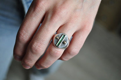 Close-up of a hand wearing a silver ring with a green stone on a blurred background
