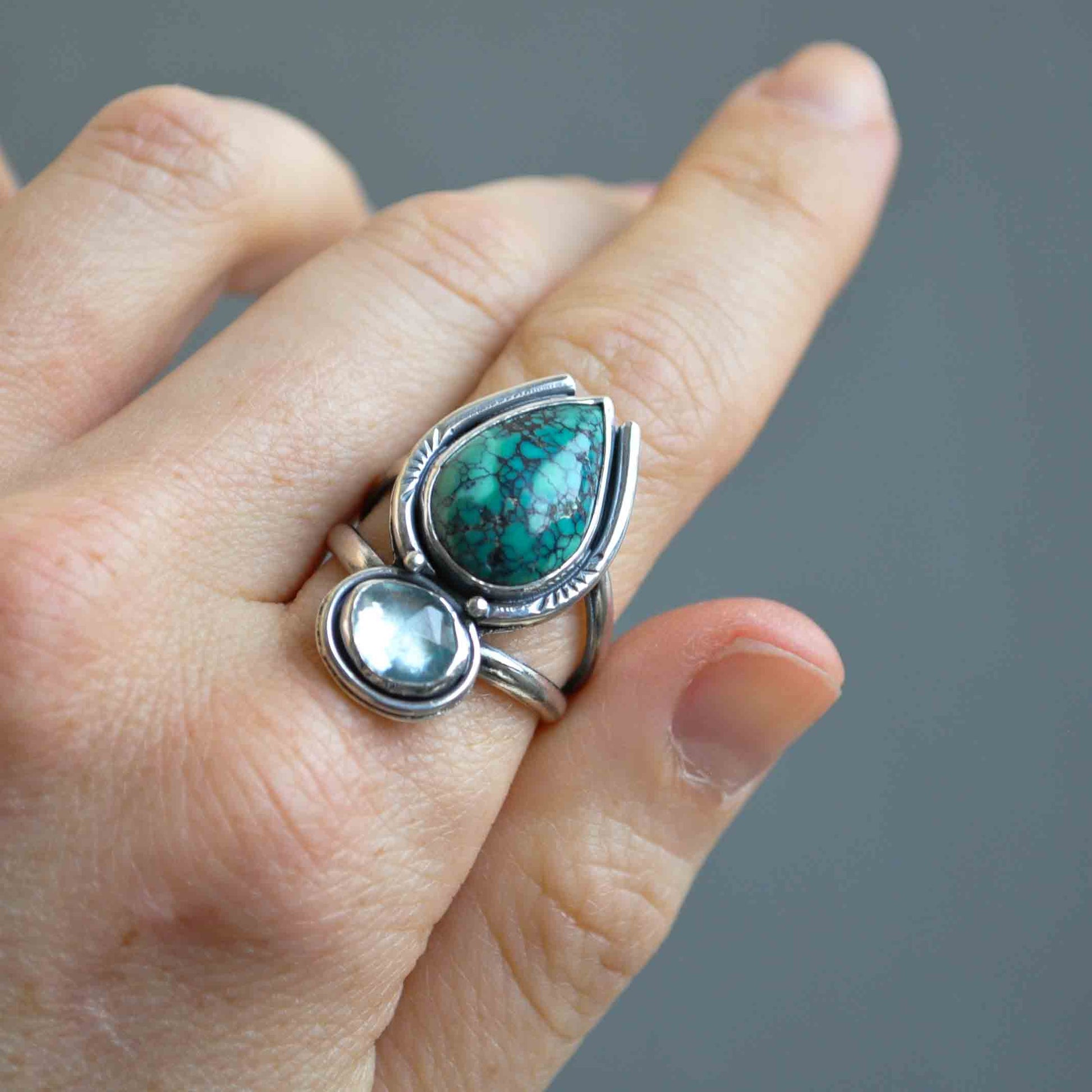 Silver ring with green and clear gemstones on a finger against a gray background