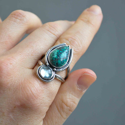 Silver ring with green and clear gemstones on a finger against a gray background